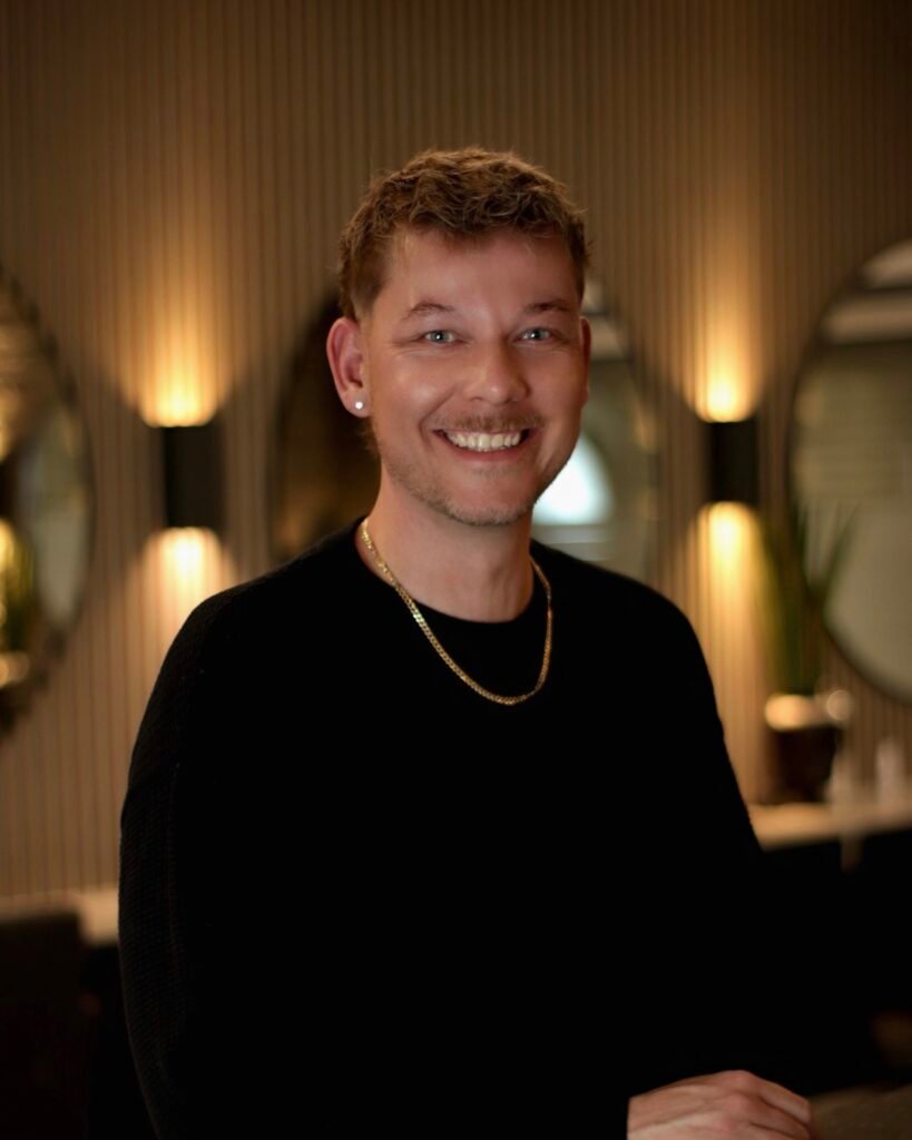 Eine lächelnde Person in schwarzem Oberteil und goldener Kette vor stilvollem, warm beleuchtetem Hintergrund.