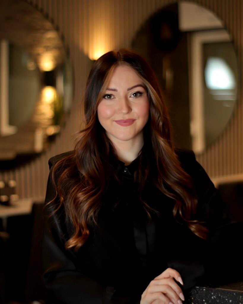 Eine lächelnde Frau mit braunem, welligem Haar in elegantem schwarzem Oberteil vor unscharfem Hintergrund.