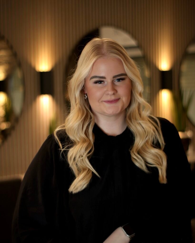 Eine Person mit langen, blonden Haaren lächelt in die Kamera; im Hintergrund warme Beleuchtung und Spiegel.