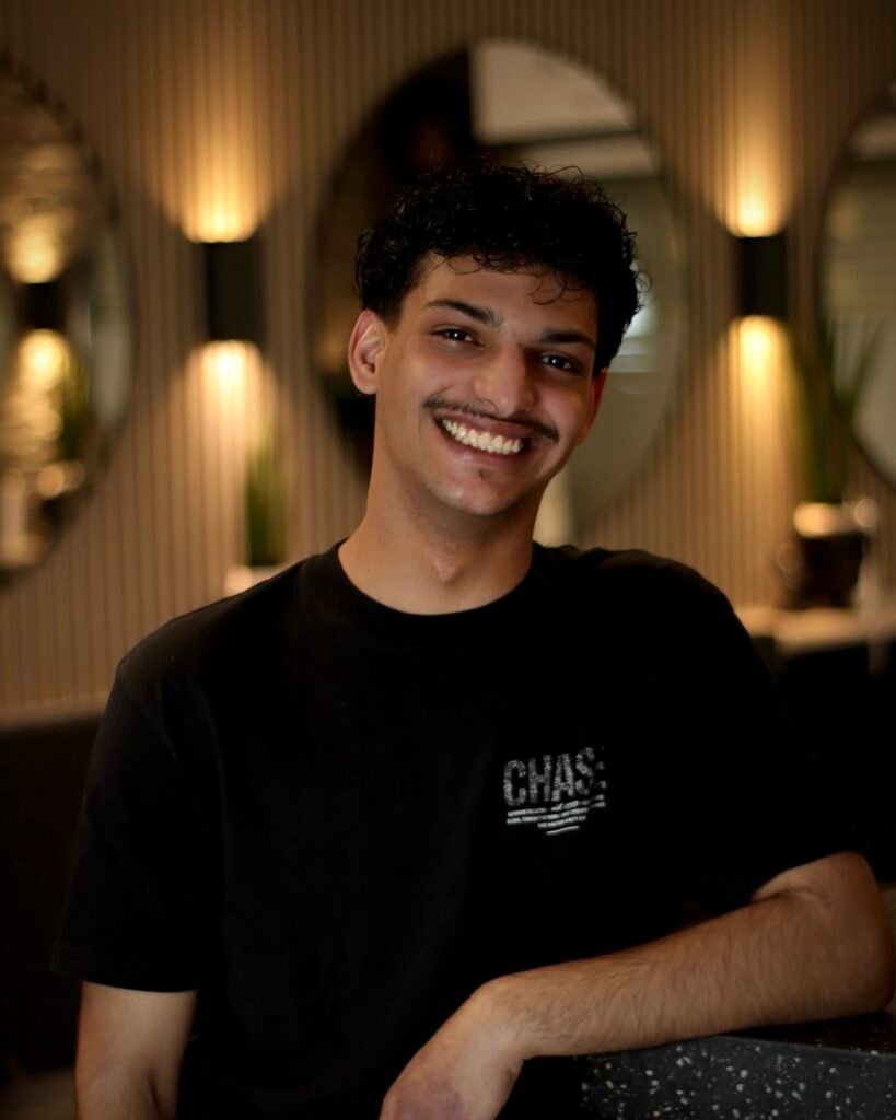 Eine lächelnde Person mit lockigem Haar und Schnurrbart trägt ein "CHASE"-Shirt in einem stilvollen Raum.