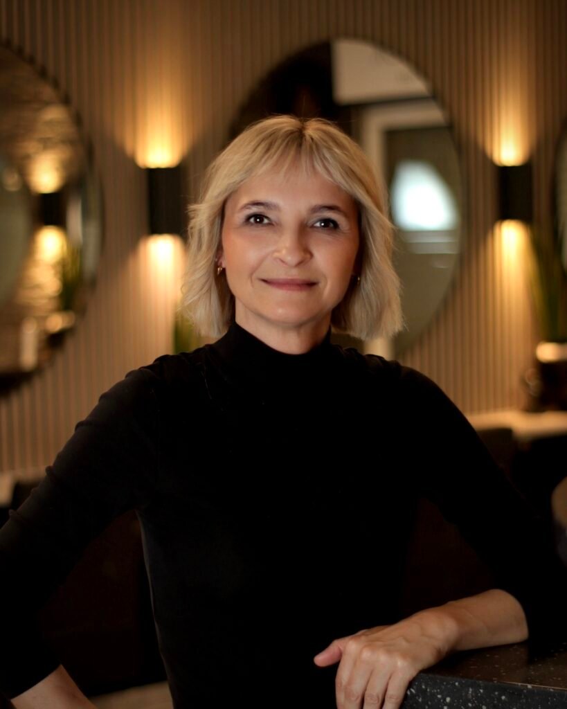 Eine lächelnde Frau in schwarzem Oberteil lehnt sich in einem warm beleuchteten, stilvollen Innenraum auf eine Theke.